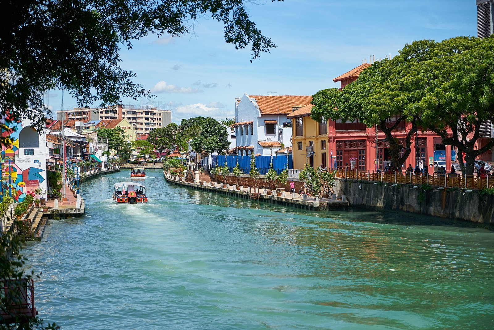 A river cruise boat on the Melaka River lined with trees and colourful buildings